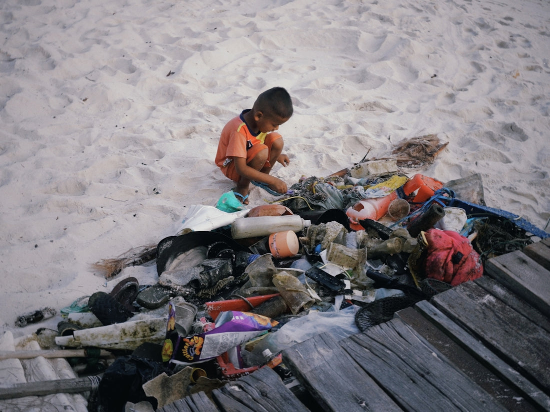 A young boy sits on a sandy beach with debris.